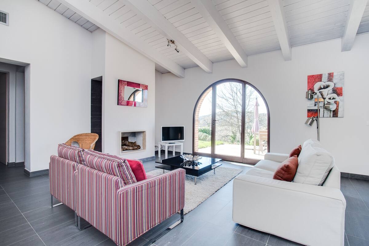 A modern living room with a striped sofa, a white sofa, a fireplace, a TV, and a large window.
