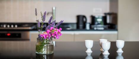 Una cocina con un florero de vidrio con flores moradas y tazas blancas sobre una superficie reflectante.