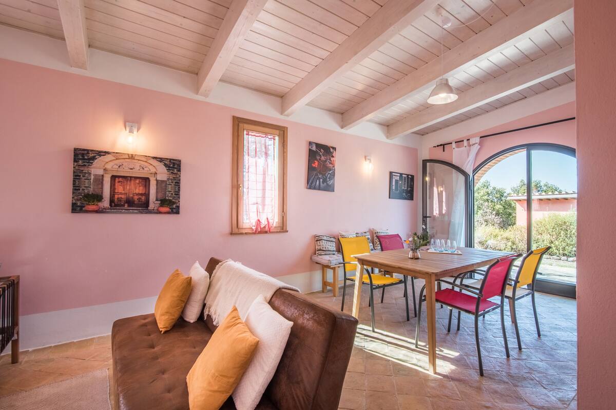 A cozy room with a brown leather sofa, a wooden dining table with colorful chairs, a large arched window, and a stone fireplace.