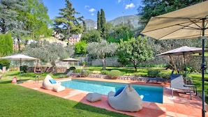A swimming pool with lounge chairs and umbrellas, surrounded by a well-maintained garden and trees. - Villa Schlosser Lake Front & Private Pool (Malcesine)