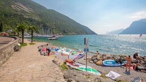 A coastal area with a stone pathway leading to a beach with boats and a mountainous backdrop. - Villa Schlosser Lake Front & Private Pool (Malcesine)
