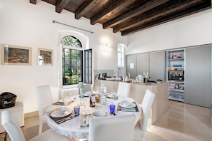A dining area with a round table set for four, white chairs, a kitchen in the background, and a window with a view of greenery. - Villa Schlosser Lake Front & Private Pool (Malcesine)