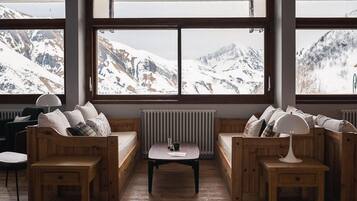 Een gezellige kamer met houten banken en een tafel, grote ramen met uitzicht op een besneeuwde berg.