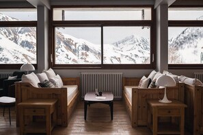 Una habitación acogedora con bancos de madera y una mesa, ventanas amplias con vistas a una montaña nevada.
