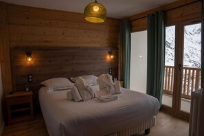 A bedroom with a wooden headboard, a bed with white linens, two bedside lamps, and a view of snowy mountains through a sliding door. - Les Cedres - Hotel, Restaurant & Spa (Saint-Sorlin-d'Arves)