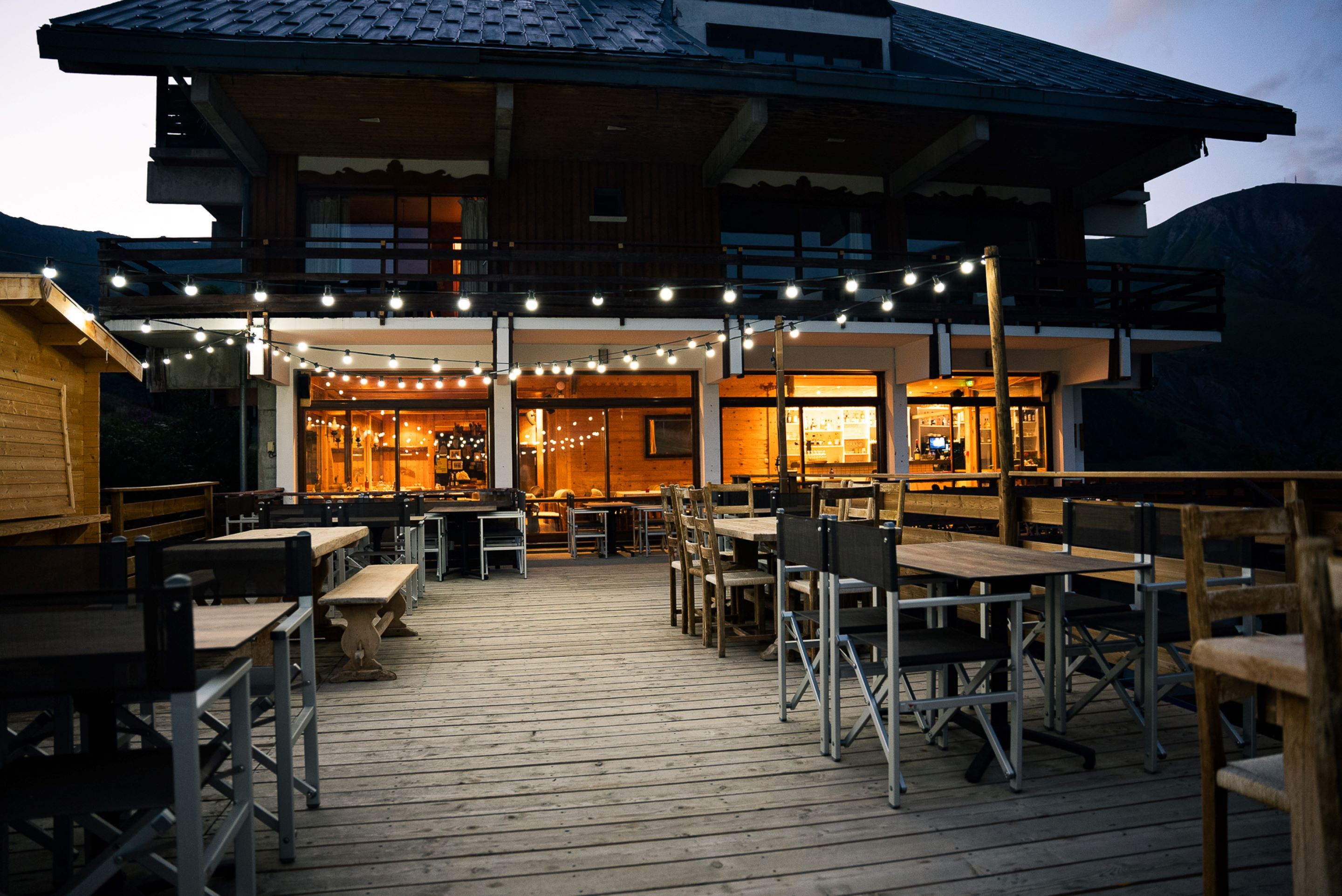 Una terrazza esterna in legno con tavoli e sedie, luci decorative a tenda e un edificio con rivestimento in legno e ampie finestre.