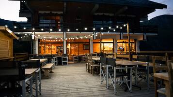 Una terrazza esterna in legno con tavoli e sedie, luci decorative a tenda e un edificio con rivestimento in legno e ampie finestre.