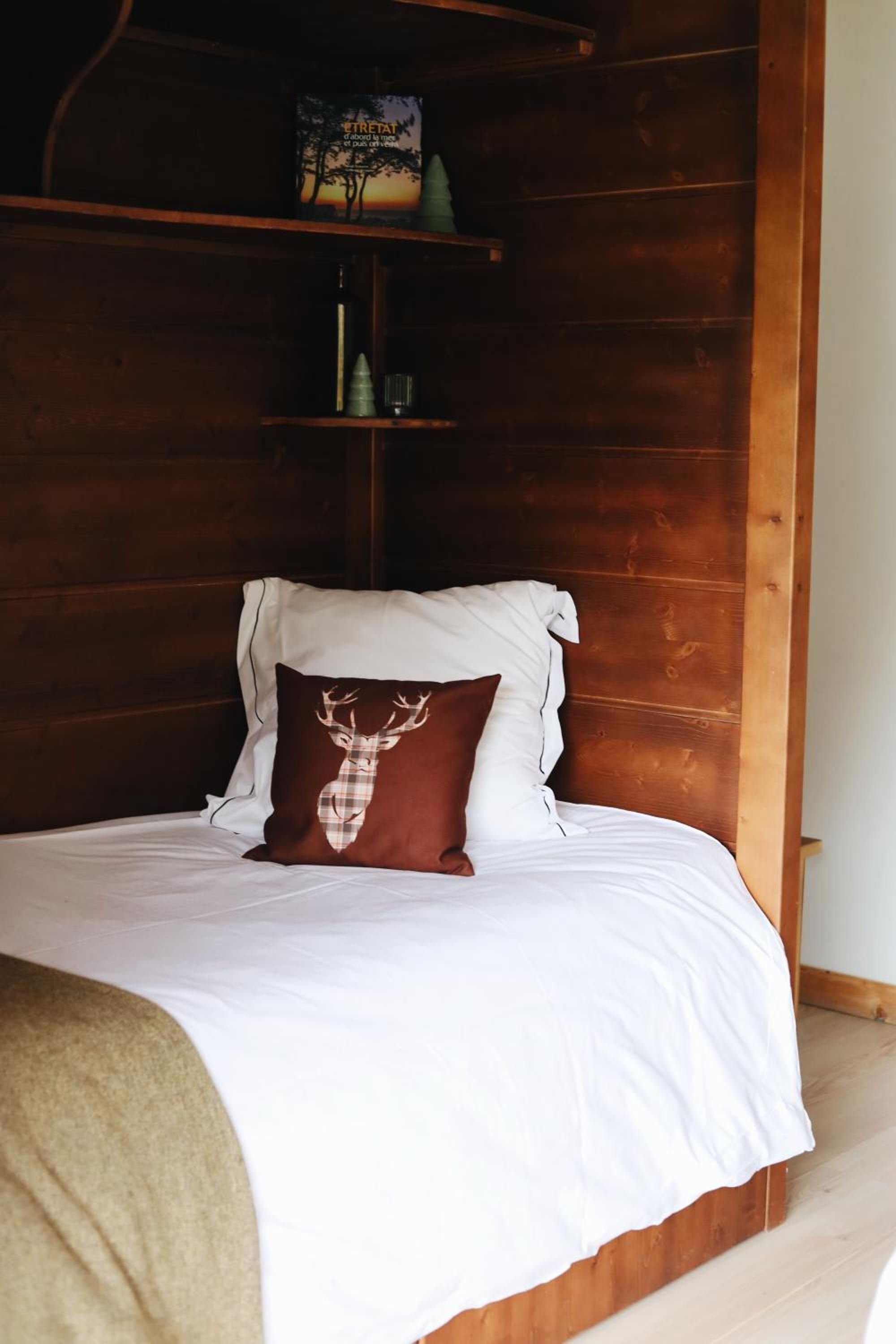 Una cama con ropa de cama blanca, un cojín marrón con un diseño de ciervo, un cabecero de madera y una estantería con un cuadro enmarcado y una botella.