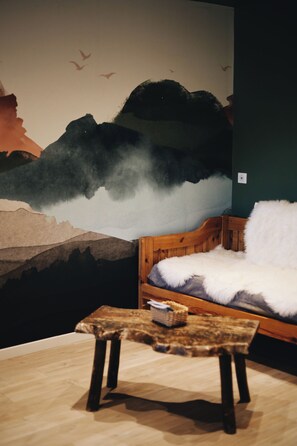 Ein Zimmer mit einem Holzbett, einem kleinen Tisch mit einem Korb und einem Wandgemälde, das eine Berglandschaft mit Vögeln zeigt.