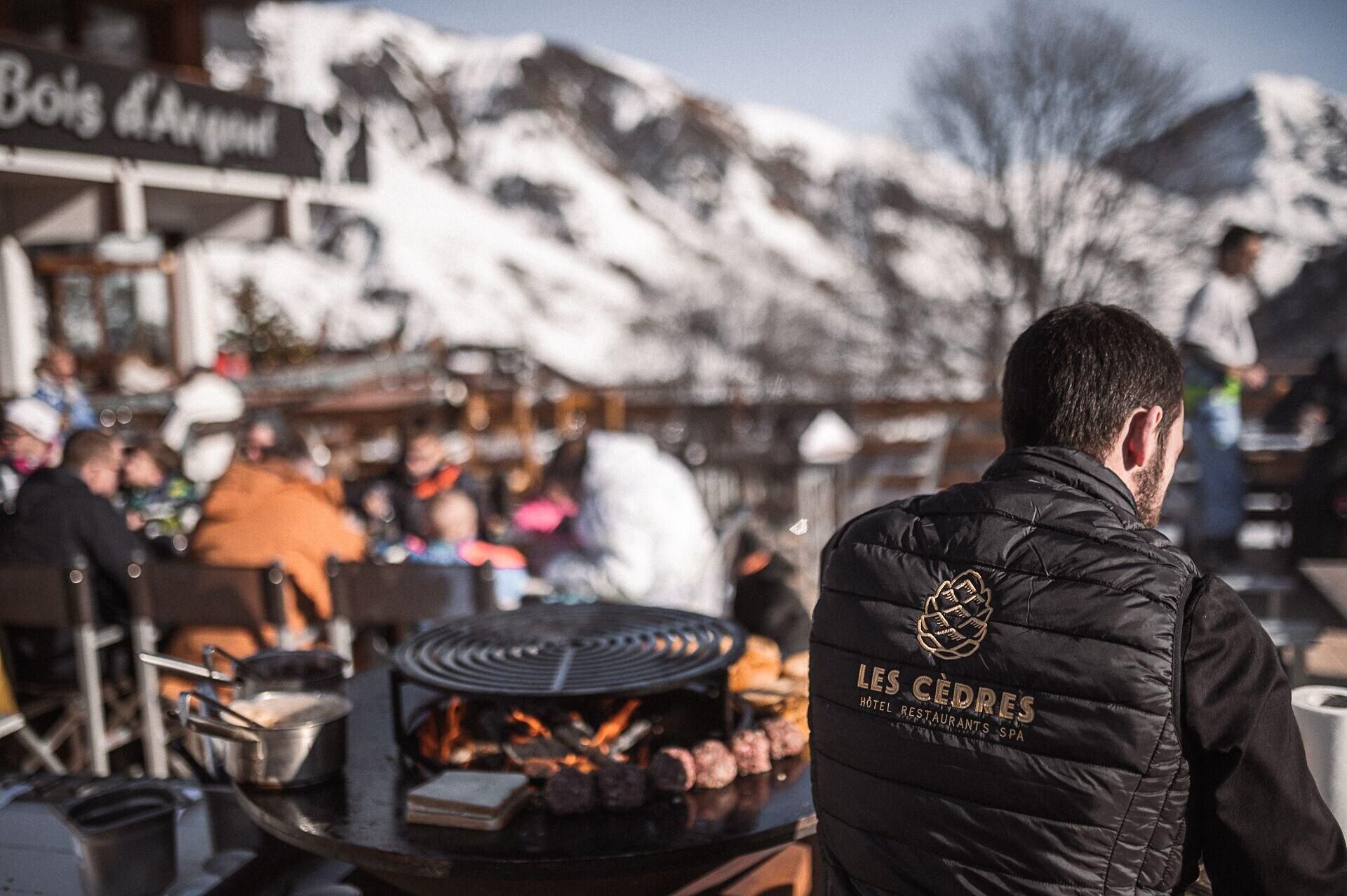 En person i en svart väst med texten ”LES CÉDRES HOTEL RESTAURANTS SPA” står framför en grill där mat tillagas.