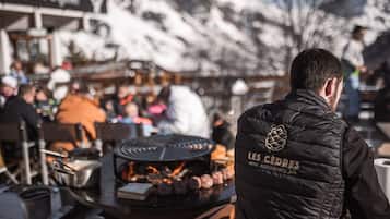 Een persoon in een zwart vest met de tekst 'LES CÉDRES HOTEL RESTAURANTS SPA', staat voor een grill waar eten wordt bereid.