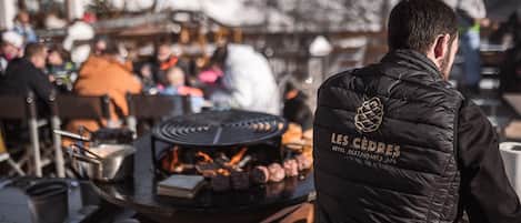 A person in a black vest with "LES CÉDRES HOTEL RESTAURANTS SPA" text, standing in front of a grill with food being cooked.