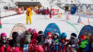 Un groupe d’enfants assis dans un télésiège, équipés de matériel de ski, entourés de montagnes enneigées et d  ’installations de ski.