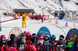 Een groep kinderen zitten op een skilift, omringd door besneeuwde bergen en skifaciliteiten, met ski's en andere ski-uitrusting.