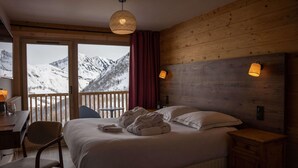 Une chambre à coucher avec un lit, un bureau, une chaise et une vue sur des montagnes enneigées, visible à travers de grandes fenêtres.