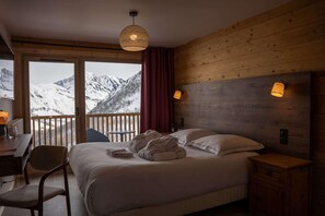 Une chambre à coucher avec un lit, un bureau, une chaise et une vue sur des montagnes enneigées, visible à travers de grandes fenêtres.