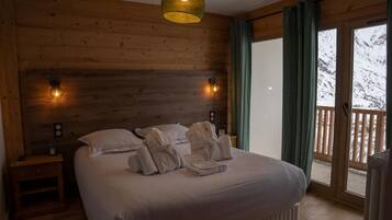 Een slaapkamer met een houten hoofdbord, een bed met witte lakens, twee bedlampen en uitzicht op besneeuwde bergen door een schuifdeur.