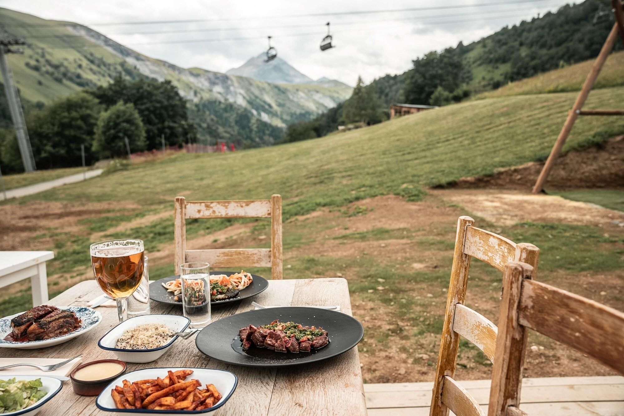 A table set outdoors with plates of food, glasses of beer, and a view of a mountainous landscape with a ski lift in the background.