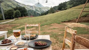 A table set outdoors with plates of food, glasses of beer, and a view of a mountainous landscape with a ski lift in the background.