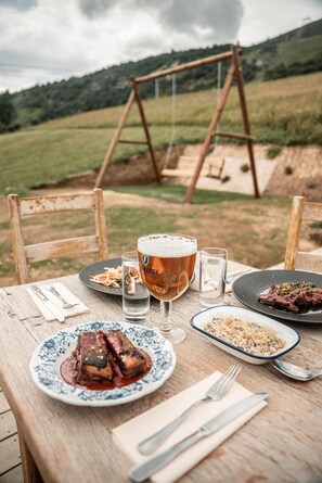 Mesa de madera al aire libre con platos de comida, una copa de cerveza y un columpio al fondo.