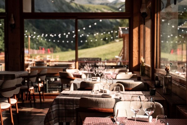 Un restaurant où les tables sont dressées pour le service, de grandes fenêtres offrant une vue sur un paysage verdoyant, et des guirlandes lumineuses au-dessus des tables.