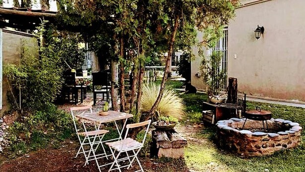 Un patio al aire libre con una mesa y sillas, un hogar para fuego y un pequeño jardín con plantas y árboles.