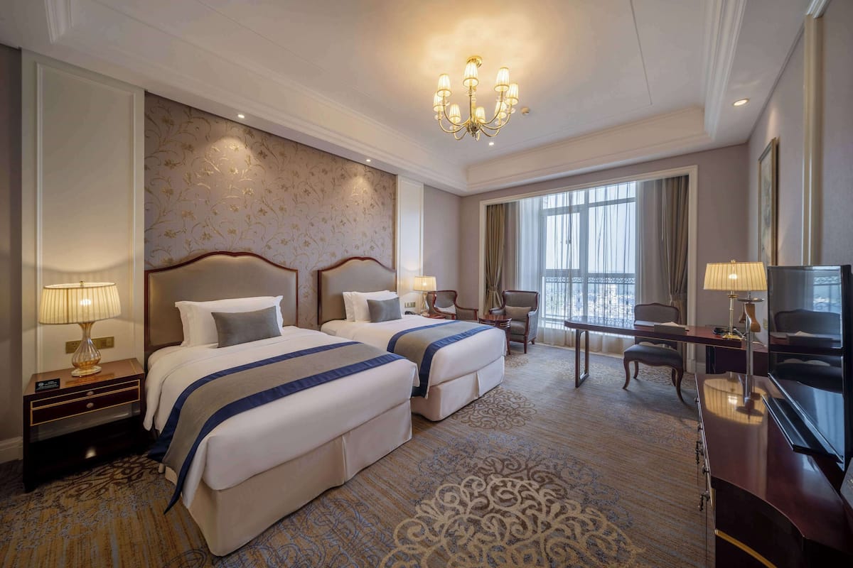 A hotel room with two beds, a desk, a TV, and a large window.