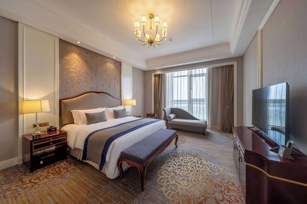 A hotel room with a large bed, a desk with a TV, a chair, and a window with curtains.