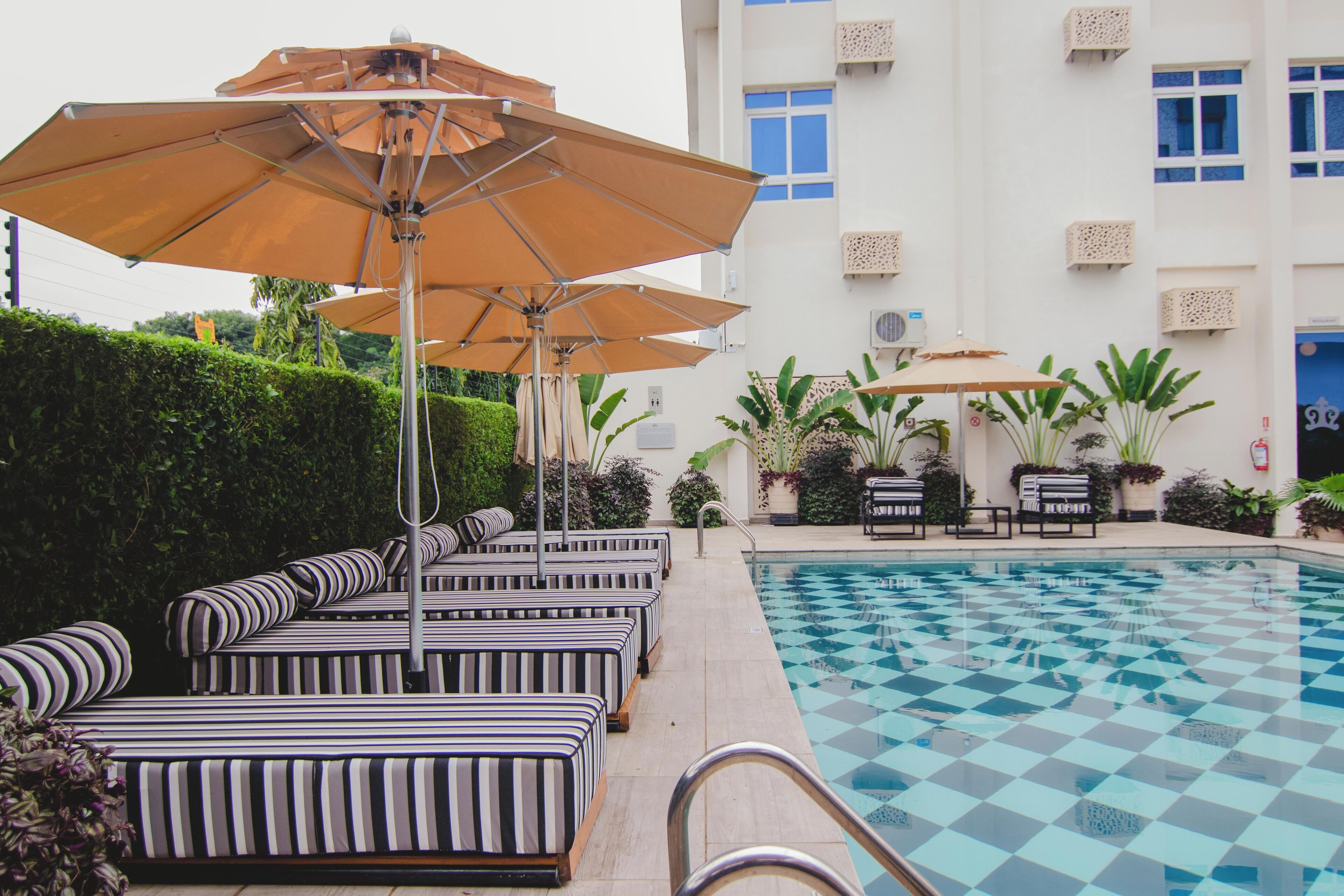 A pool area with striped lounge chairs and umbrellas, surrounded by a building with multiple windows and air conditioning units.