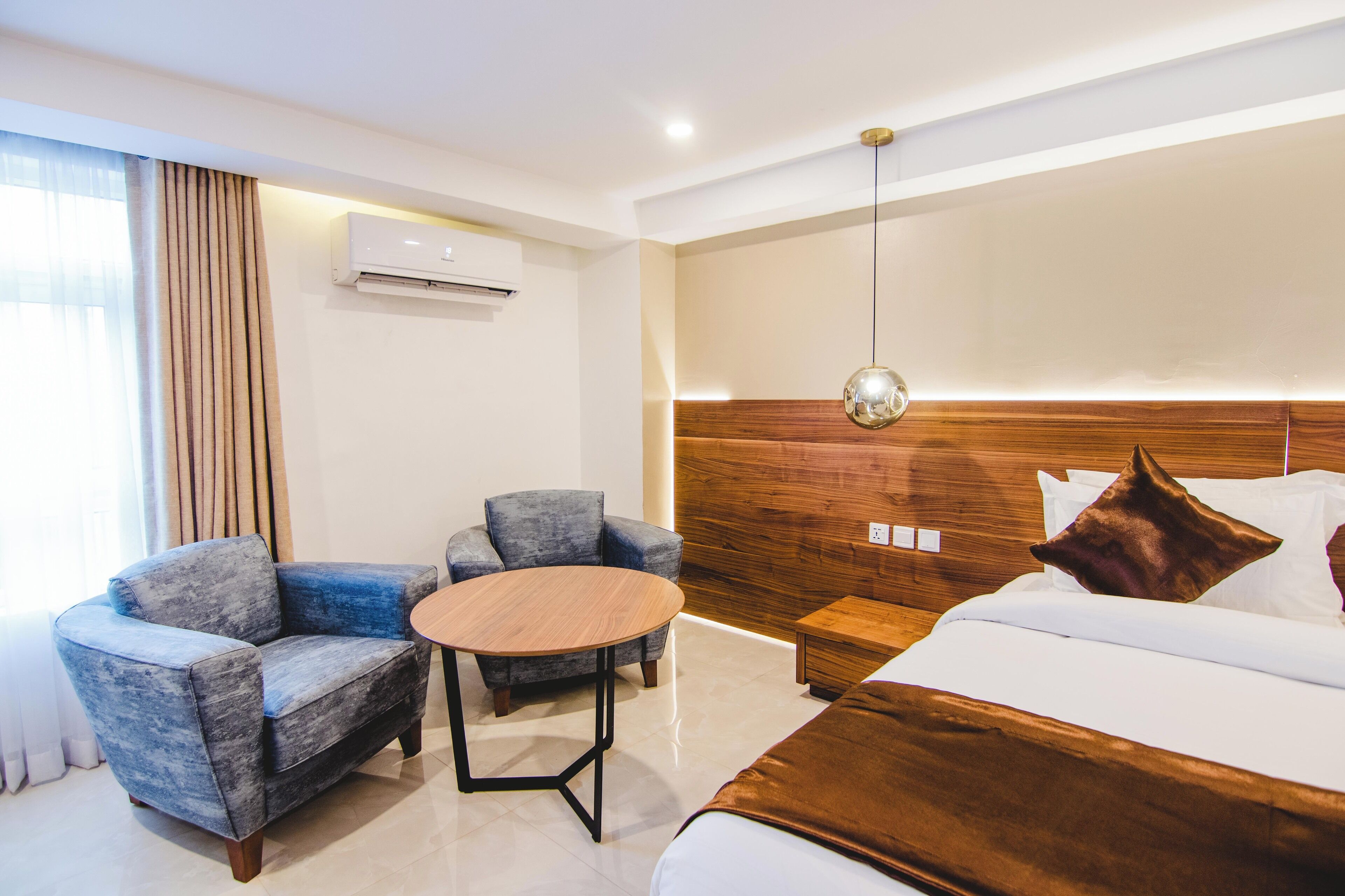 A modern hotel room with a bed, two armchairs, a small round table, a wooden headboard, and an air conditioning unit.
