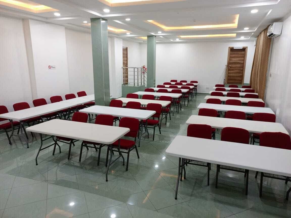 Une salle de conférence avec des rangées de tables blanches et de chaises rouges, équipée de luminaires au plafond et de climatiseurs.
