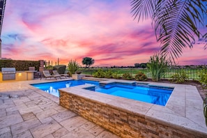 A swimming pool with a brick surround, lounge chairs, and a grill area.