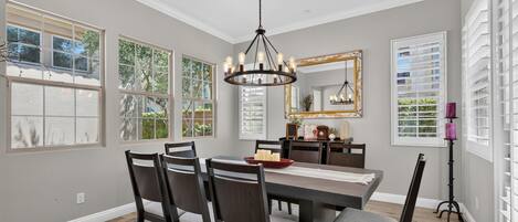 Une salle à manger avec une grande table, des chaises, un lustre, un miroir et des fenêtres.