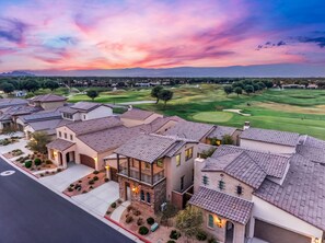 A residential area with houses featuring terracotta roofs and stucco walls, a golf course in the background, and a sunset sky.