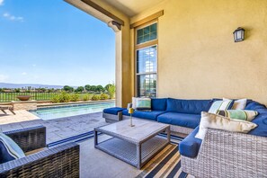 A sunlit outdoor seating area with a pool, wicker furniture, and a view of a landscaped garden.