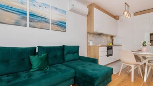 A modern living room with a green sectional sofa, a dining area with a white table and chairs, and a kitchen with white cabinets and wooden accents.