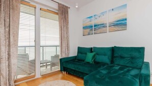 A teal sofa with green cushions, a beige rug, and a view of the sea through a window with blinds.