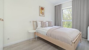 A minimalist bedroom with a wooden bed, a small white side table, a window with curtains, and a wall-mounted artwork.