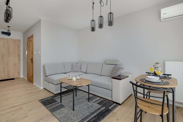 A modern living room with a gray sectional sofa, wooden flooring, and a dining area with pendant lighting.
