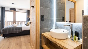 Une chambre d’hôtel moderne avec un grand lit, une tête de lit en bois, une grande fenêtre avec des rideaux, une salle de bain avec un lavabo blanc et un miroir.