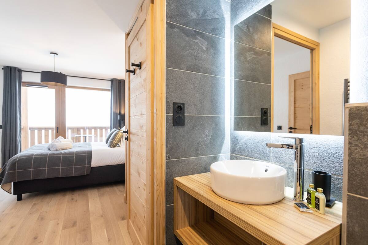Ein modernes Hotelzimmer mit einem großen Bett, einem Holz-Kopfteil, einem großen Fenster mit Vorhängen sowie einem Badezimmer mit Waschbecken und einem Spiegel.