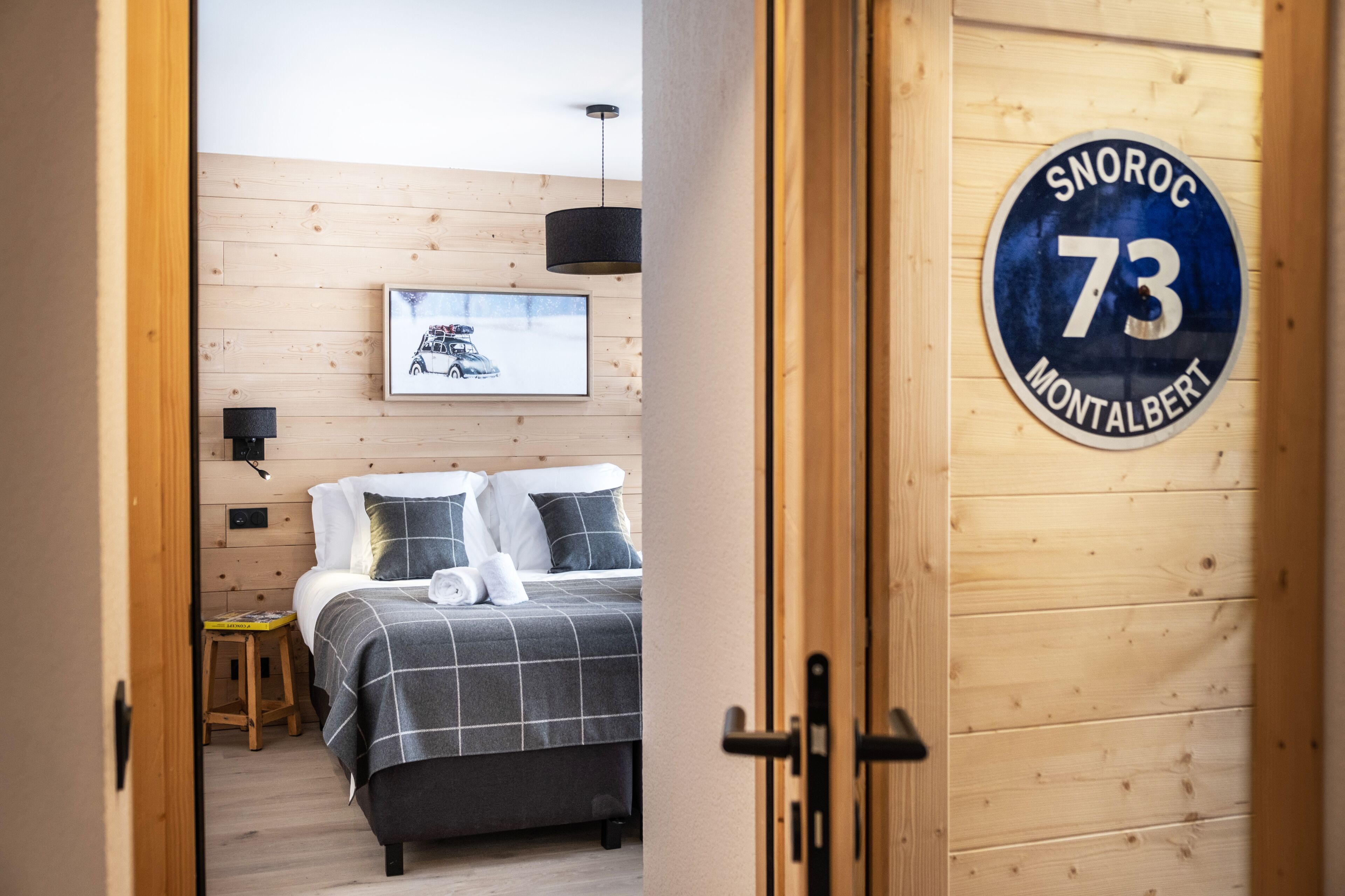 Une chambre d’hôtel avec un lit, un tableau encadré au mur et une porte sur laquelle est apposée une plaque indiquant « SNOROC 73 MONTALBERT ».