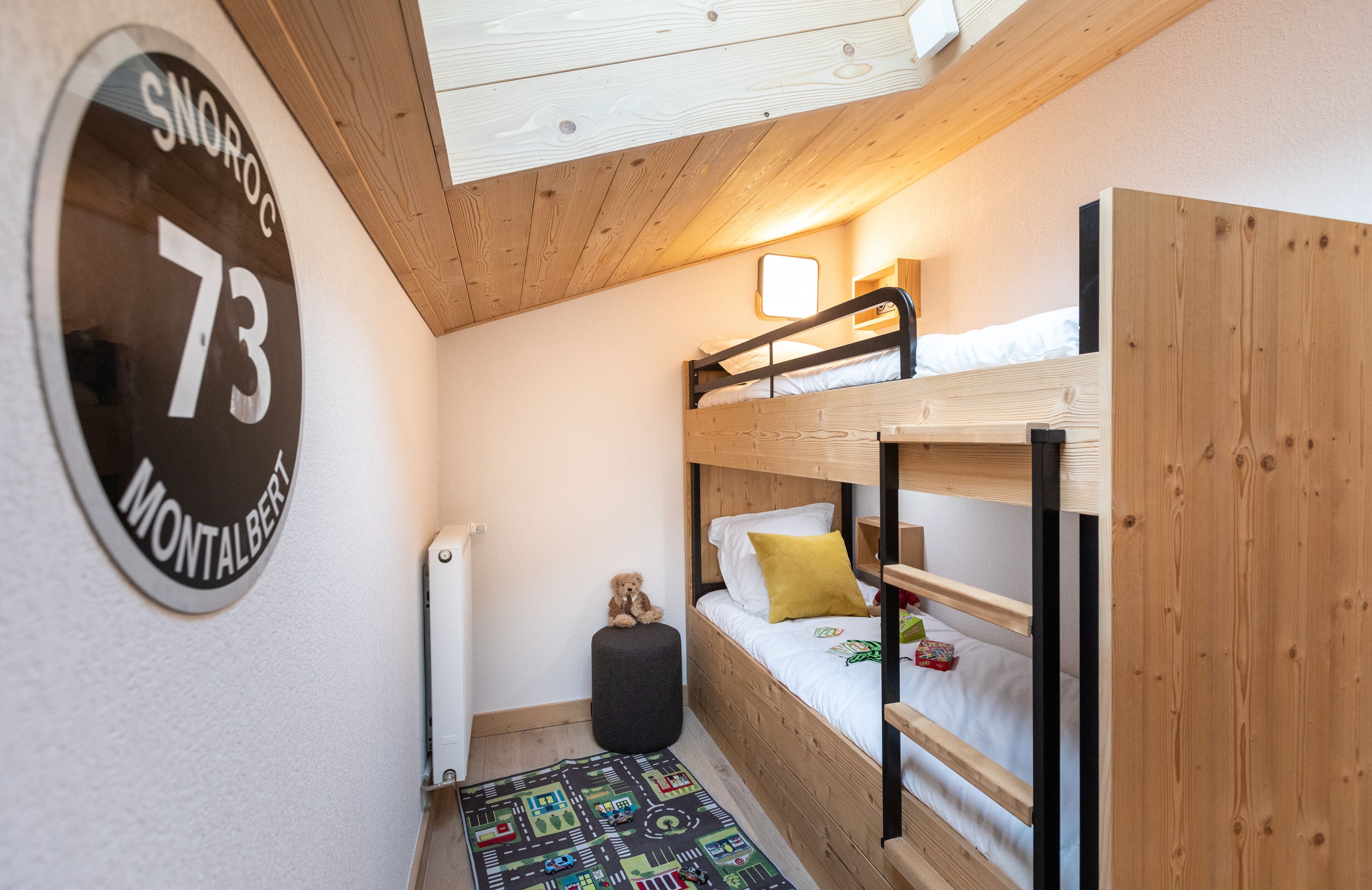 Habitación con literas, techo de madera, un reloj redondo en la pared y un tapete de juego colorido.