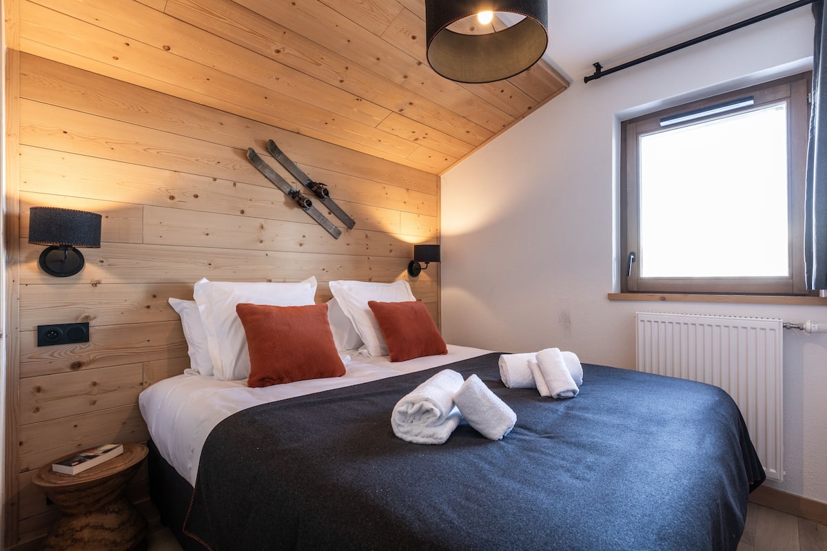 Ein Schlafzimmer mit Holzboden, einem großen Fenster, einem Bett mit weißen und roten Kissen sowie einer schwarzen Bettdecke.