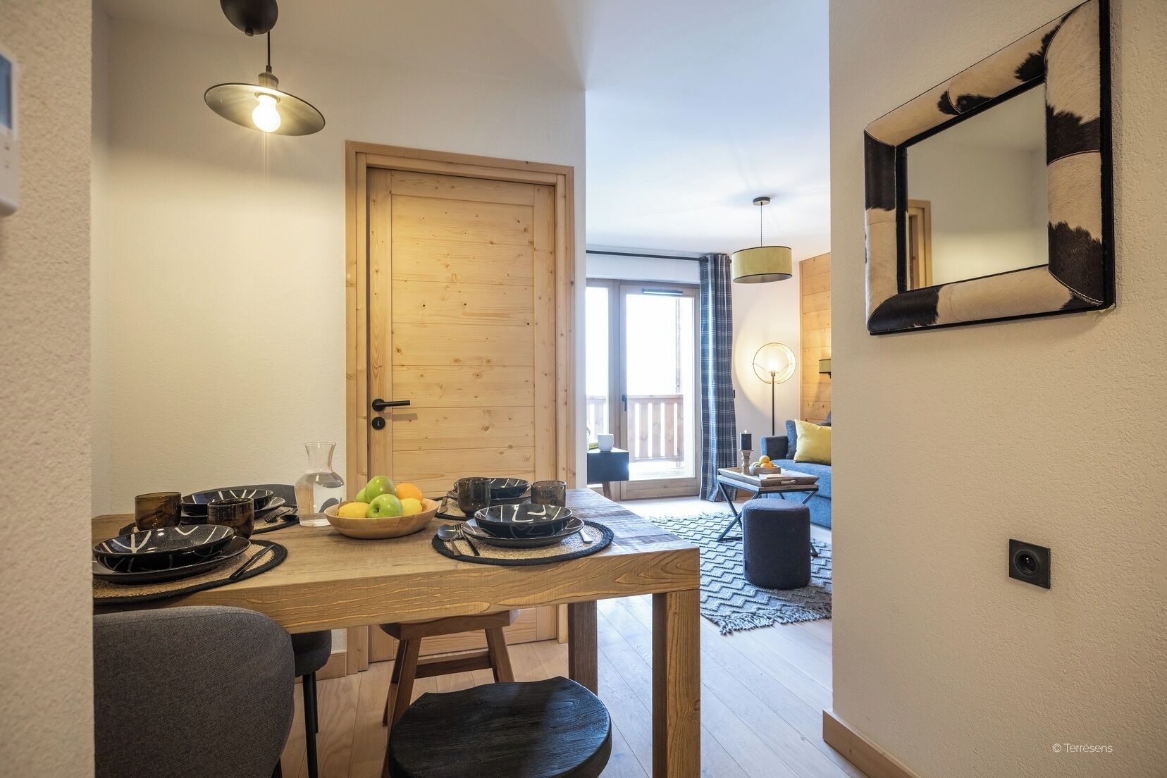 Een eethoek met een houten tafel voor vier personen, een schaal fruit en uitzicht op het balkon door een houten deur.