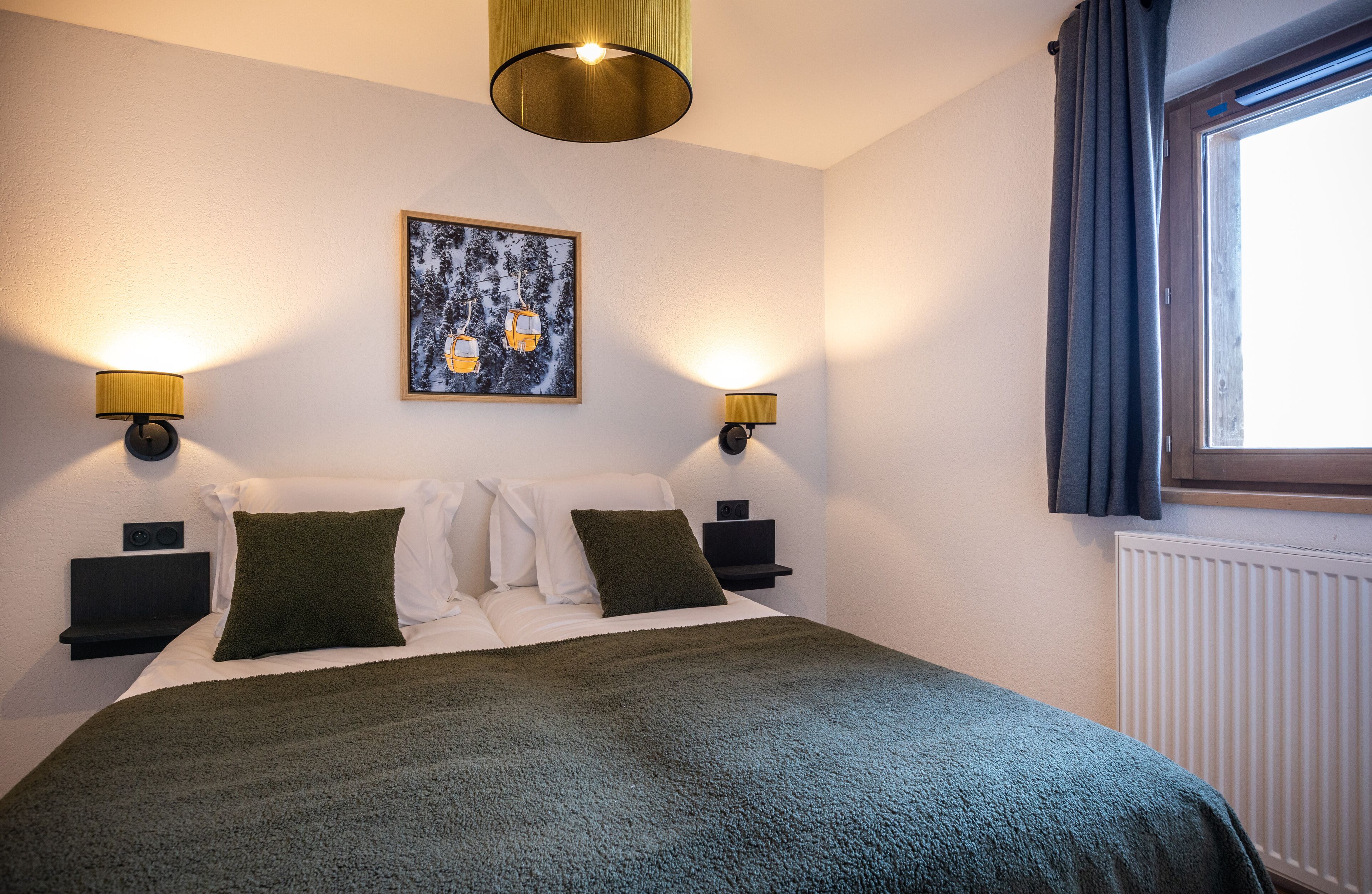 A neatly made bed with a green blanket, two bedside tables with lamps, a framed picture on the wall, and a window with curtains.