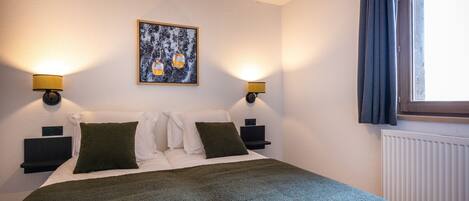 A neatly made bed with a green blanket, two bedside tables with lamps, a framed picture on the wall, and a window with curtains.