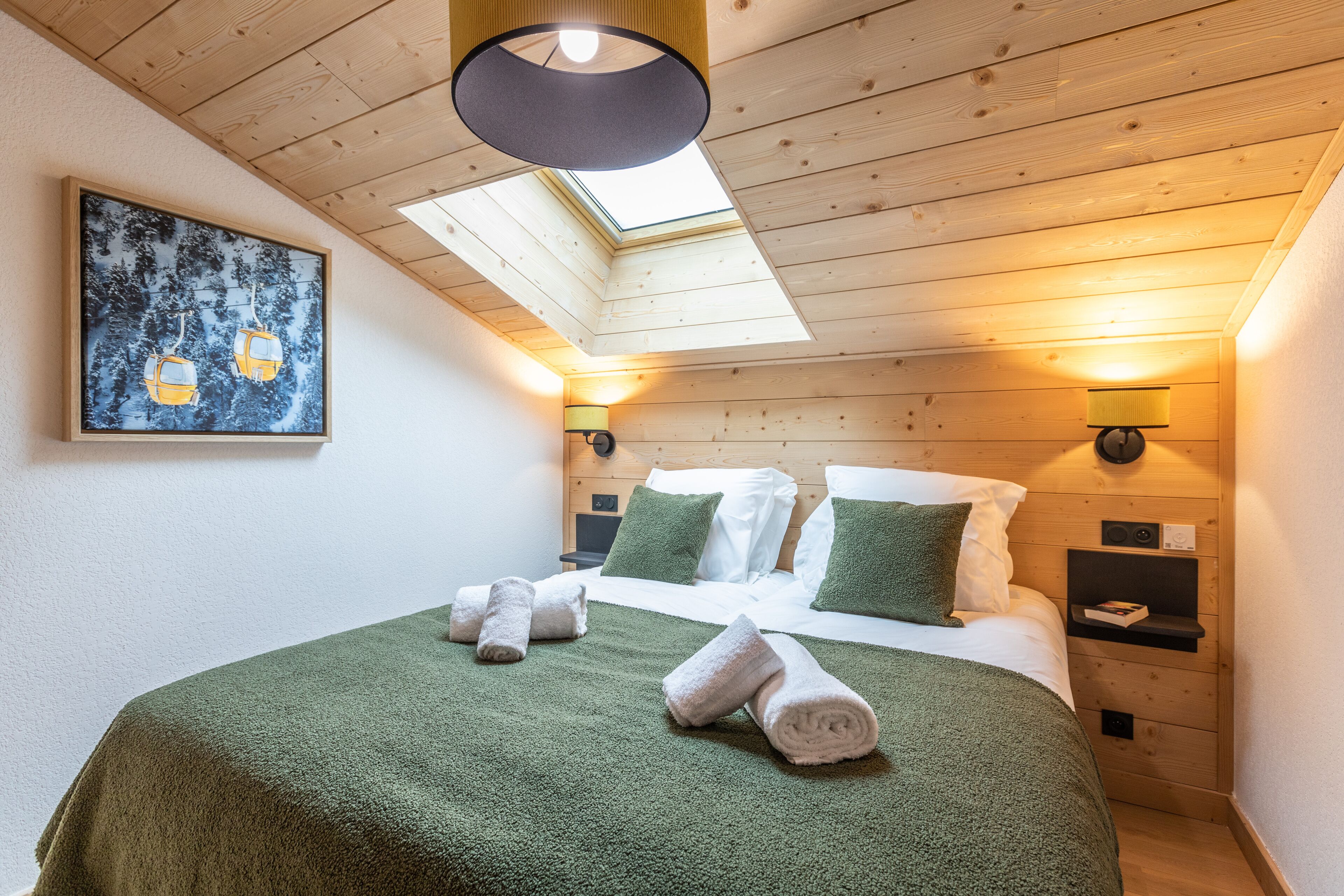 A hotel room with a bed, green towels, a painting of a ski lift, a skylight, and a nightstand with a lamp.