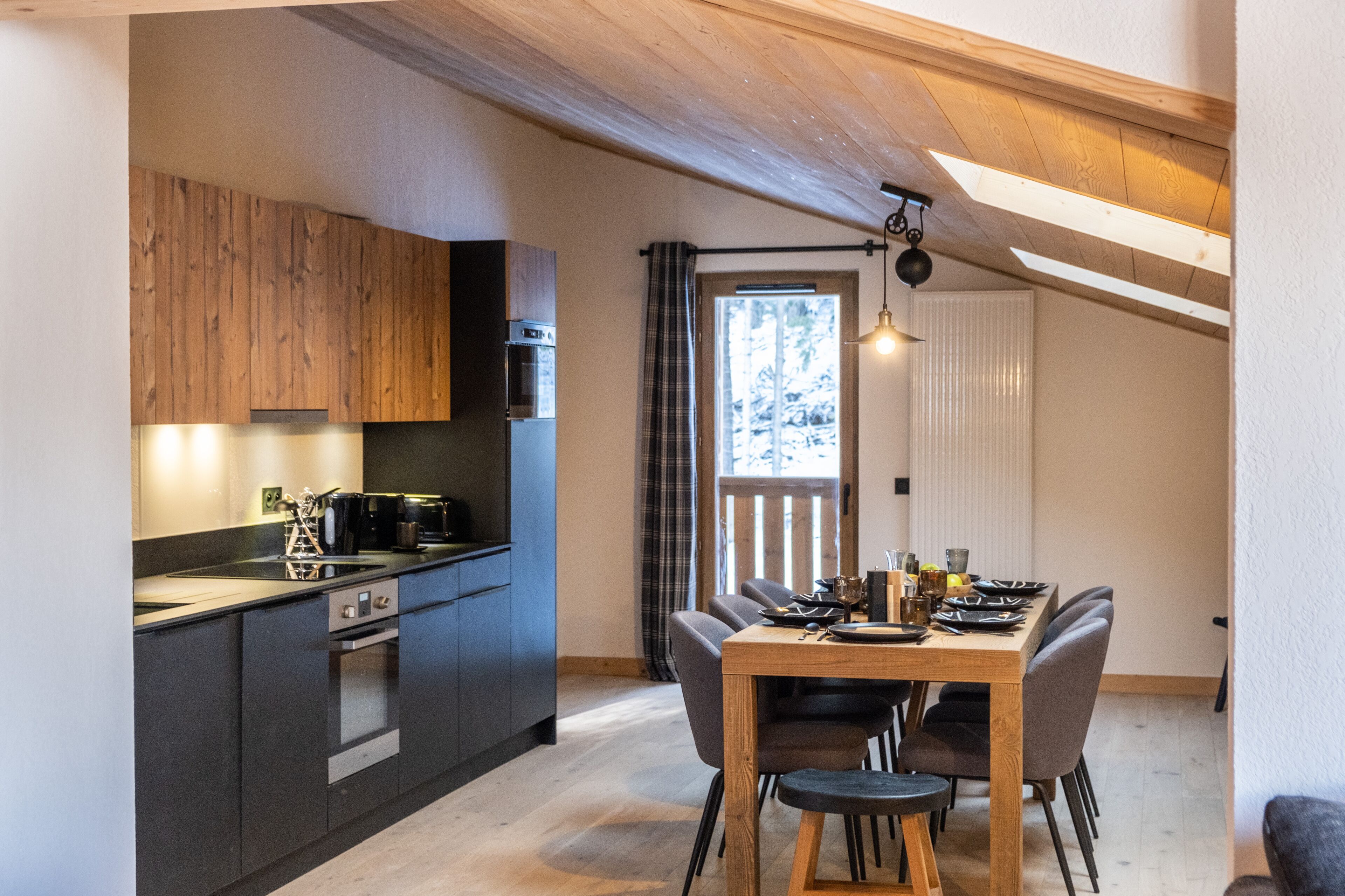 A modern kitchen with dark cabinets, a wooden dining table, and a view of the outdoors through a window.