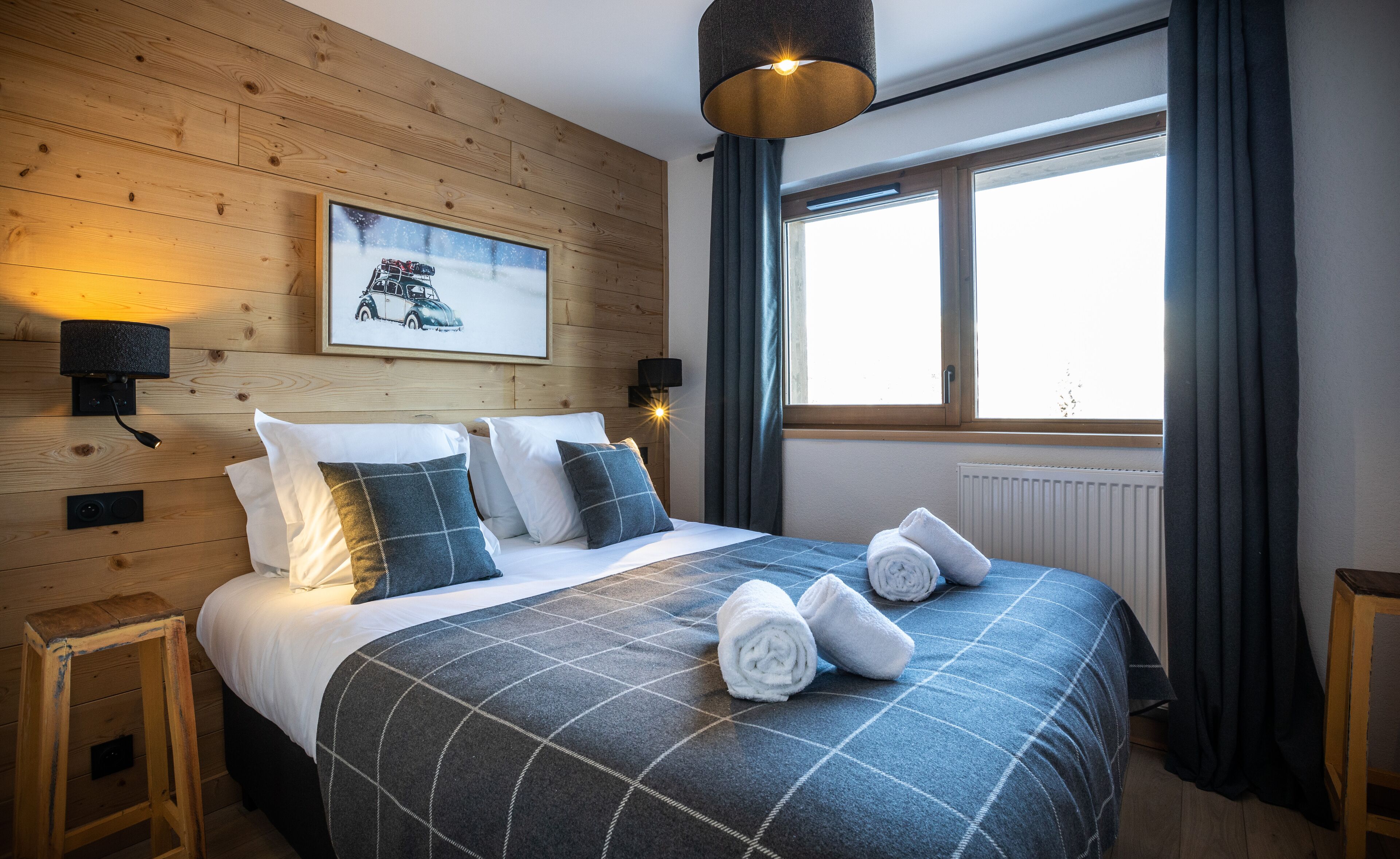 A neatly made bed with a checkered duvet, two wall-mounted lamps, a framed picture of a car, and a window with curtains.